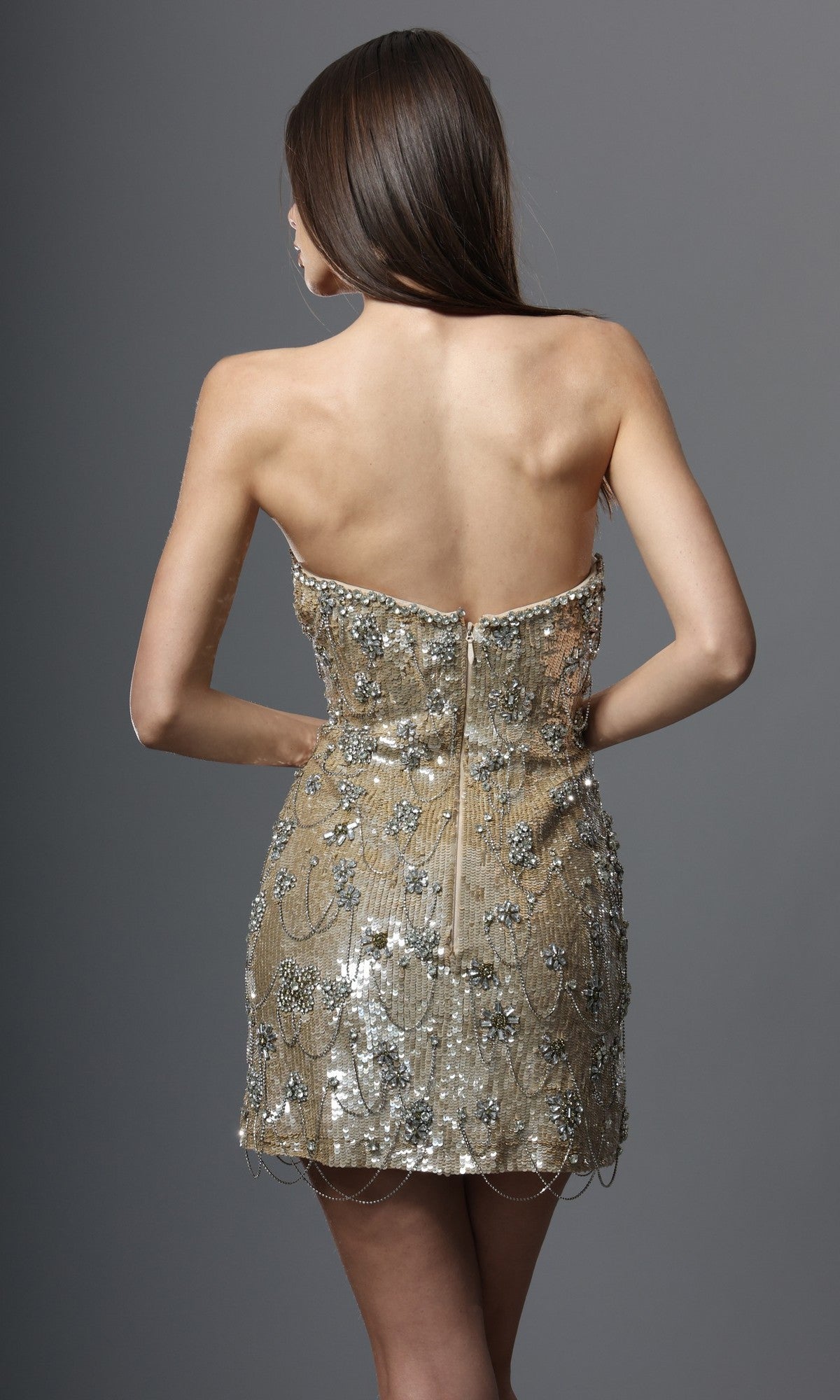 A woman stands with her back to the camera, wearing the Jovani 52529 short homecoming dress—an elegant, strapless designer mini dress embellished with gold and silver sequins in intricate floral patterns—set against a plain gray background.